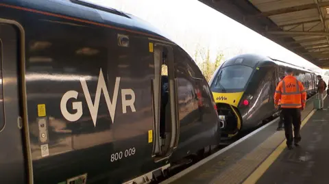 Two GWR trains at a railway station platform, with passengers waiting and a railway worker in a high-visibility orange jacket walking along the platform.