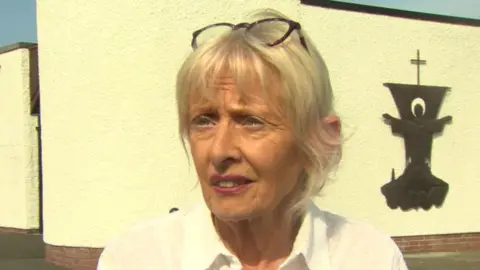 Nuala is standing outside the chapel wearing a white linen shirt. She has whispy white/blonde short hair and her tortoise shell glasses are on her head. 