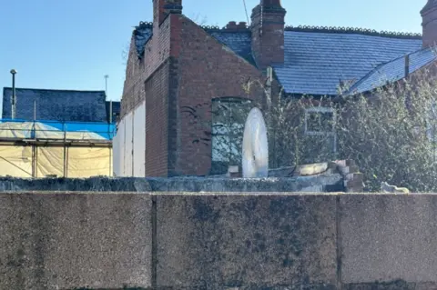 A view from behind the properties, showing that demolition work has taken place. A circular saw is seen in a block of concrete, with bricks to the side of it. 