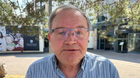 A man with short grey hair slightly smiles at the camera. He wears glasses and a glue and grey striped shirt and is sitting outside 