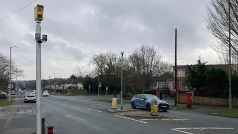 A small yellow camera on top of a large metal pole by the side of a road