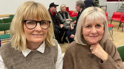 Diane Heggie and Rachel Wilcox look at the camera shoulder to shoulder. They are both wearing brown coloured jumpers. Diane has a white collar.