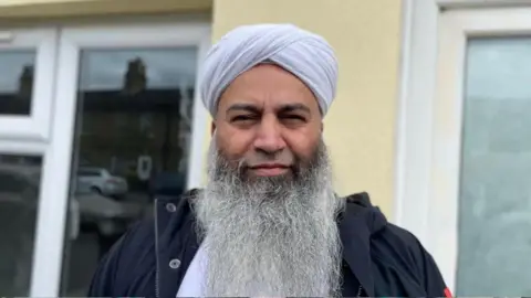 Headshot of Mohammed Saeed looking at the camera. He is wearing a white turban and a dark coloured jacket.