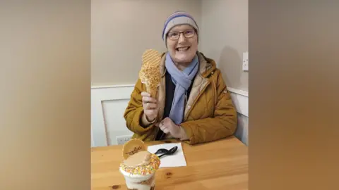 Christine Campbell A woman, Christine Campbell. She is sat inside, smiling, wearing a mustard yellow coat, a purple scarf, a stripey woolly hat and glasses. She is holding an ice cream on a cone with a circle wafer slice in it and multicoloured sprinkles. In front of her on the table is a napkin with cutlery on it and a tub of ice cream with multicoloured sprinkles, sauce and a circle wafer slice on top. 