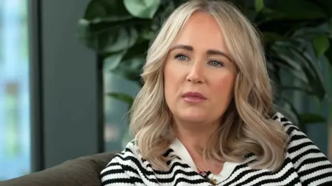 Joanne Dorrian, a woman with shoulder-length blonde hair and wearing a black and white striped top with a gold necklace, looks off camera.