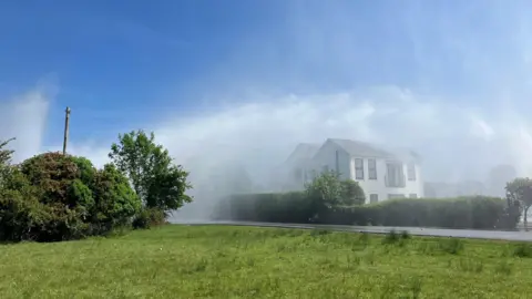 A huge spout of water can be seen spurting over a couple of white detached houses. There is a large spray of water hanging in the air as mist.