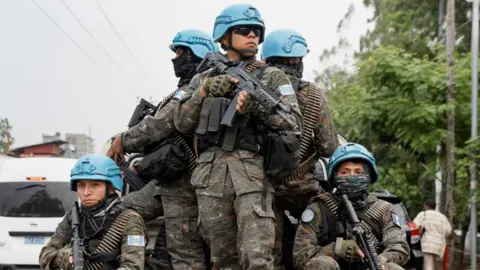 Five soldiers wearing blue hats stand on the back of a truck in front of trees in Goma, DR Congo.