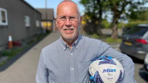 Annan Athletic Man standing with football under his arm. He has thinning grey hair and glasses and is in a blue and white checked shirt. 