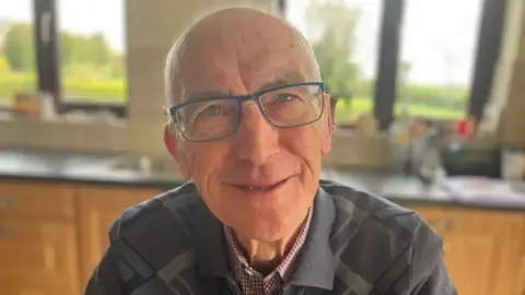 Alphie Lonergan. He is bald, wearing a grey jumper with shirt underneath.