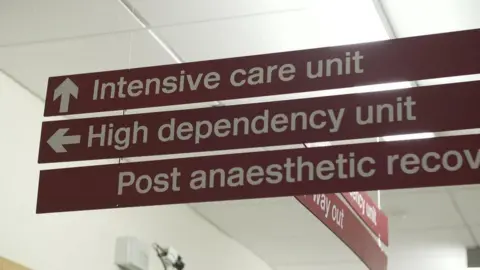 A sign in a hospital ward. It is red, with white lettering, an up arrow is directing people tpo intensive care, a left arrow to a high dependency unit.