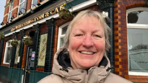A woman with short blonde hair wearing a beige coloured coat, standing outside a pub.