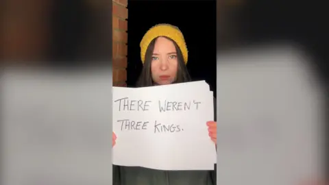 A woman in a yellow beanie hat holds up a piece of paper with the words 'There weren't three kings'