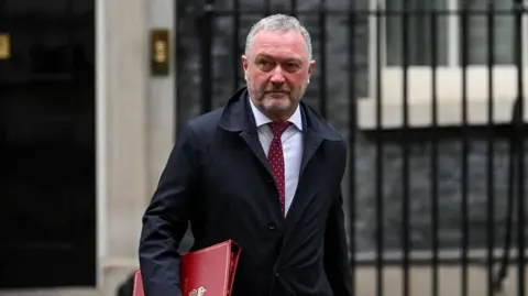 Reuters Steve Reed in a dark overcoat, white shirt and red tie walks outside a government building, carrying a large red document folder under one arm. The background features stone walls, a doorway and black metal railings. The photograph is taken at street level with the person in sharp focus and the building softly blurred behind.