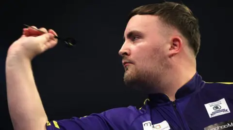 Getty Images Luke Littler is aiming a dart in his right hand, he is wearing a purple top with sponsors on it, the background is black.