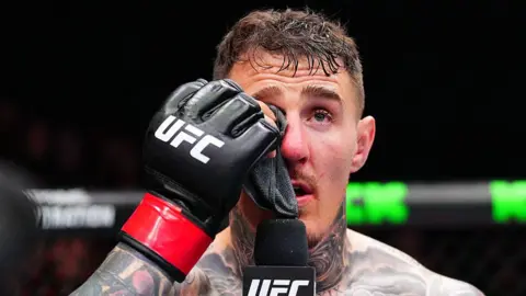 Tom Aspinall holds a cloth to his eye and speaks into a microphone