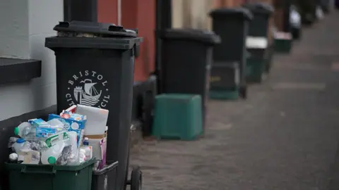 Getty Images Close-up of black wheelie bins labelled “Bristol City Council” standing on a street, alongside smaller green recycling bins, with house doorways visible in the background.