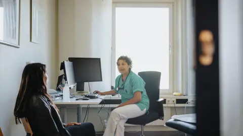 A medical worker sat with a patient in a consultant room. The consultant has black hair and is wearing a green shirt with white trousers and sat near a computer. 