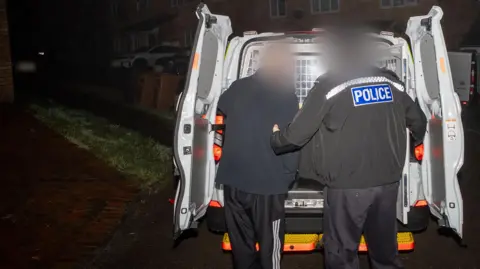 Environment Agency A police officer leads a suspect into the back of an open police van, parked on the pavement of a housing estate. Both their faces have been blurred. It is dark. 