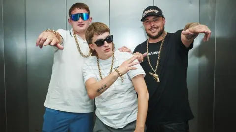 Three young men pose in gold chains and sunglasses. They appear to be standing in a metal lift