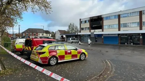 Police cars and ambulance vehicles are parked by the small block of flats with one flat clearly fire damaged and blackened with windows and their framework blown out