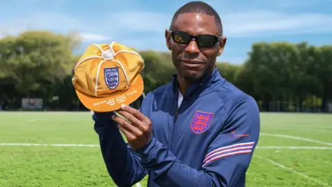 Darren Harris A man with short hair, sunglasses and wearing a blue England team jacket, smiles as he stands on a football pitch and holds a yellow cap with an England badge on the front.
