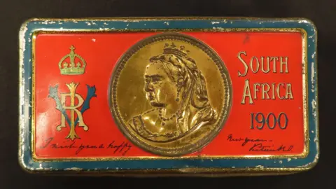 Lockdales Auctioneers and Valuers A tin box. It is red on the top with a gold picture of Queen Victoria and the words "South Africa 1900".