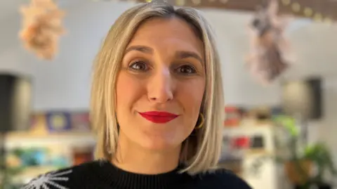 A lady with blonde hair cut in a short bob, she has brown eyes and is wearing red lipstick and gold hoop earrings. She is looking directly at the camera and smiling, and is wearing a black jumper with stars on it.