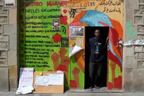 AFP Ugandan Francis Kashamba poses at the frontdoor of the abandoned school of La Massana, in the center of Barcelona, on July 3, 2018, occupied since mid-April by dozens of migrants. Once far from the strife that pushed them to come to Europe, what has awaited thousands of illegal migrants in Spain is a form of agonising purgatory, according to the migrants occupying the Massana School