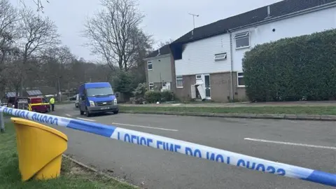 Police tape on a residential road with houses behind. One of the houses has fire damage.