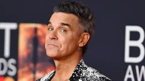 Getty Images Robbie Williams has dark hair and is wearing a fancy black and white jacket. He is smiling at the camera.