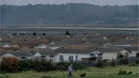A wide shot of a series of white houses with grey roofs. In front of the houses there is green land and a man wearing a purple coat walking a black dog.