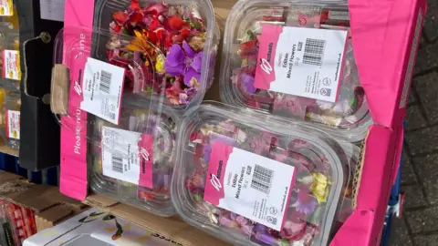 Towcester Community Larder A number of boxed edible mixed flowers, piled up in a pink cardboard box. 