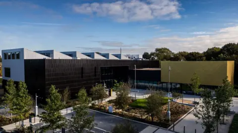 A photograph of ARU Peterborough's campus. The building is built on two sides, with a black-clad and white-clad building on the left and a black and yellow clad building on the right. In front are trees, grass, paths and car parking. 
