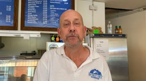 Dean Cummings at New Forest Finest Fish and Chips. He is bald, with short grey stubble, and is wearing a white polo shirt.