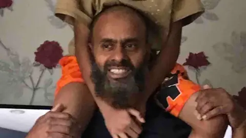 Humayra Ali Masrob Ali pictured with a child on his shoulders. He wears a t-shirt and is smiling. He appears to be standing in a living room.