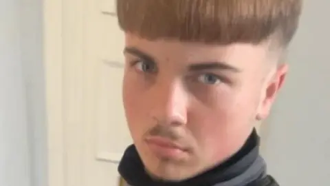 Aidan Tottman is taking a selfie with brown hair brushed to the front of his face and shaved around the side of his head. He is wearing a Nike jumper and is stood in front of a white door