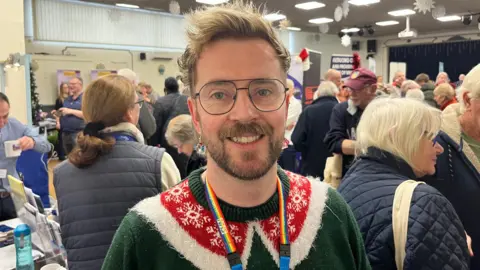 John Devine/BBC Jack Avison has light brown wavy hair in a sideways quiff on top of his head,. He has a brown moustache and beard and is wearing glasses and a dark green jumper with red and white Christmas icicle patterns on it. Behind him are lots of people in a function room.