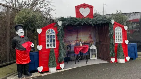 A small hut with red doors opening out each side with white windows. White hearts are adorned on them with two triangular plants. Inside is a table for two wit a red sheet and wine and glasses on it. Behind it is a Parisian draped dispaly. A statue of a chef is stood next to the hut.
