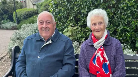 Kate Bradbrook/BBC Robert Ward with short white hair and Pamela Ward with medium-length white hair smiling at the camera. Robert has a blue jacket while Pamela has a purple sweater and red, white and blue scarf. They are sitting on a black bench in front of a hedge.
