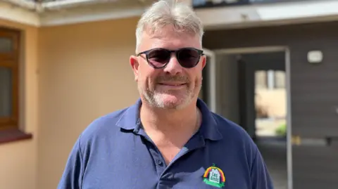 Shaun has short light hair, a beard, and sunglasses stands smiling in front of a cream‑coloured house with a tiled roof. He is wearing a blue textured polo shirt with a small colourful logo on the chest. The background shows a sunny sky and part of an adjoining building.