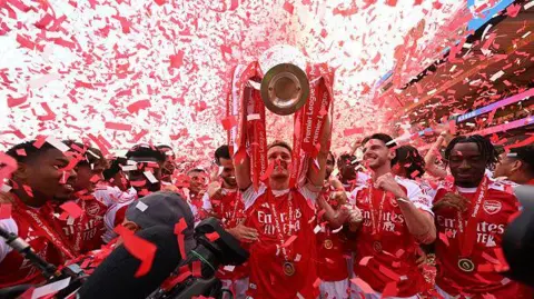  An Arsenal player lifts the Premier League trophy above his head as red and white confetti falls around him, with teammates in red shirts celebrating alongside.