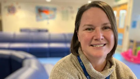 A woman is smiling at the camera. She has short dark hair and is wearing a beige jumper. She has a navy blue lanyard around her neck which has "RUHX" written on it. 