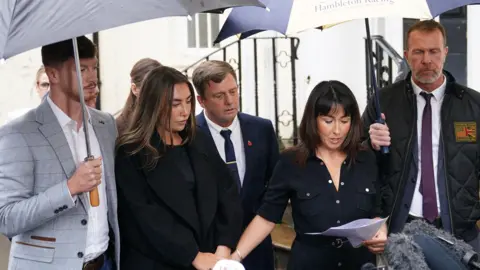 PA Media A woman wearing a black dress is holding sheets of paper and talking to the media. She is holding hands with another woman, and surrounded by a group of people.