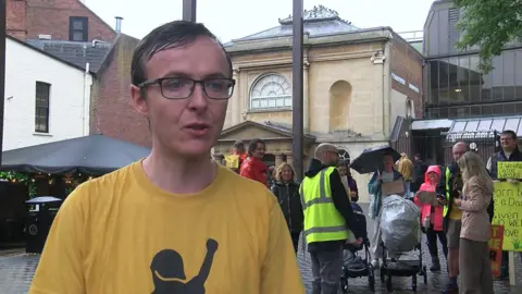 A man with glasses and a yellow shift is giving an interview. Behind him, women and men are striking for better paternity leave. They hold placards with slogans. It is a rainy day. There is a pram beside one of them.