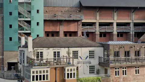 Google The Boat Club building pictured in 2018. It is white with Boat Club written in blue at the front and two paddles painted on it. The Prince Bishop multi-storey car park is behind it. The building is mostly brown brick, except for its tall staircase wall, which is turquoise. 