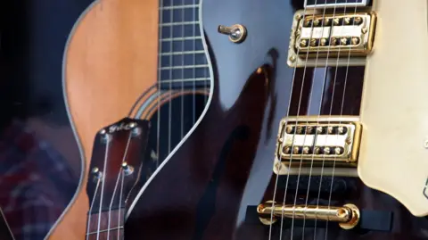 A close-up of a selection of guitars - with a wooden acoustic one in the background, a neck in the foreground and a polished dark brown and cream electric guitar at the front of the image.