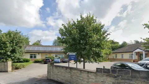 Crescent Dale in Heckmondwike. There is a sign outside the complex which consists of two single-storey buildings and a car park. There are two trees in the foreground.