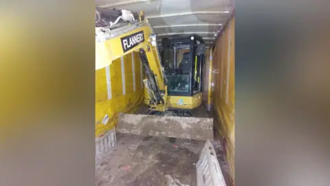 Cambridgeshire Police A yellow digger is at the back of a lorry. There is mud on the floor and a silver ladder which is lying on the floor in the bottom right corner. 