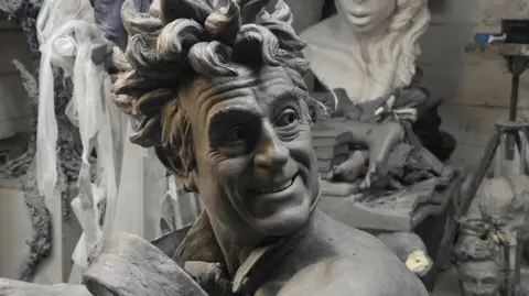 A close up picture of the clay model used for the sculpture showing Terry Jones with wild hair and smiling. He wears only a tie that looks like it is blowing in the wind