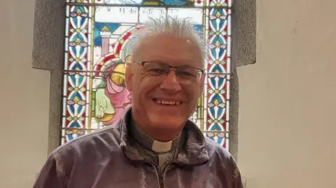 A man with spiky grey hair is pictured wearing a purple fleece with a clerical collar visible beneath. He has glasses and is smiling broadly. A stained glass window is visible behind him. 